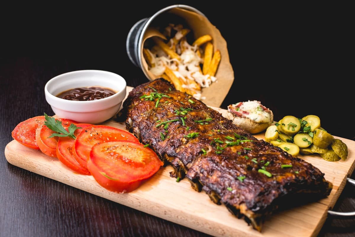 Smoked ribs on a serving board with pickles, tomatoes, and barbecue sauce against a dark background