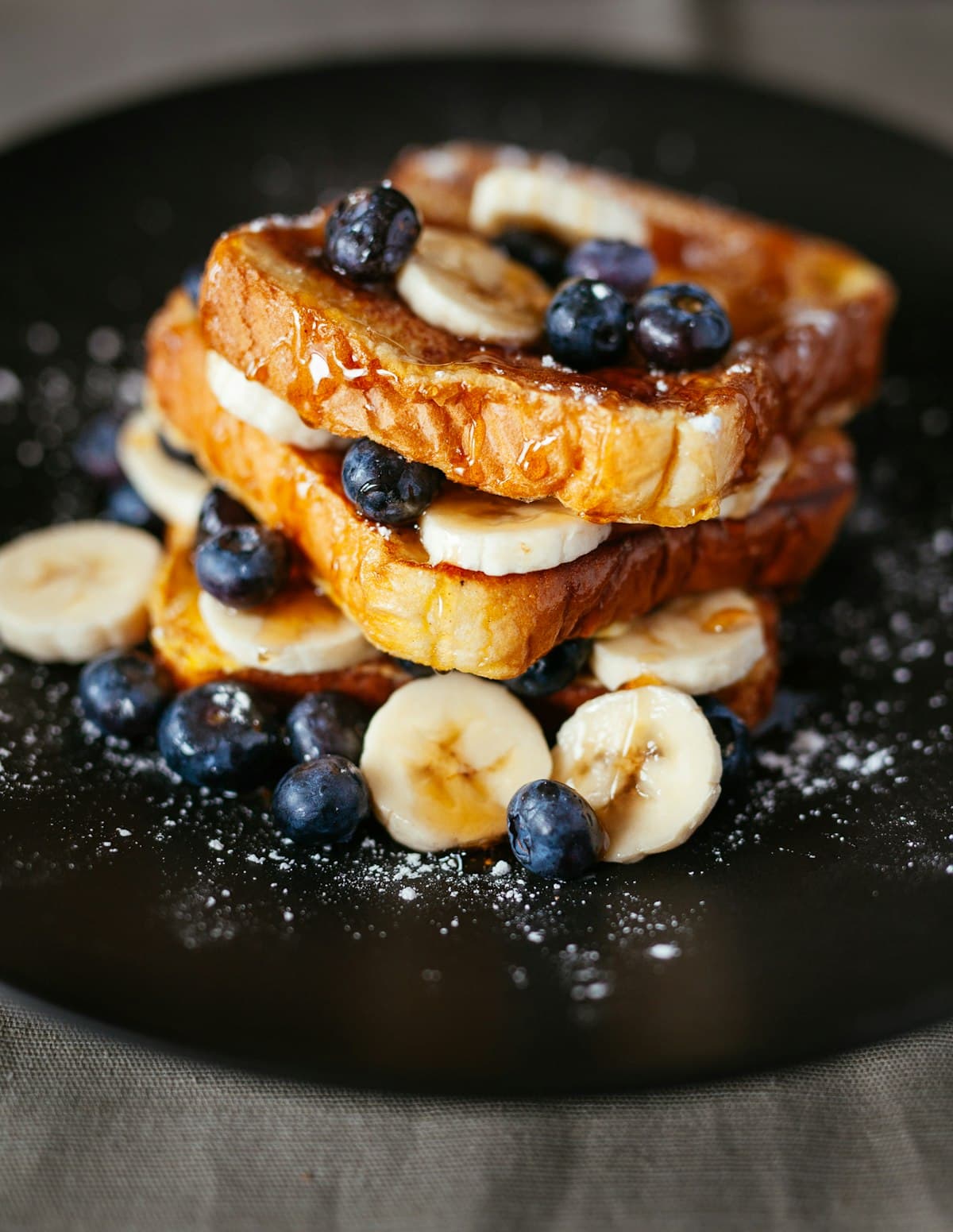 Stack of French toast topped with blueberries, banana slices, and powdered sugar on a dark plate