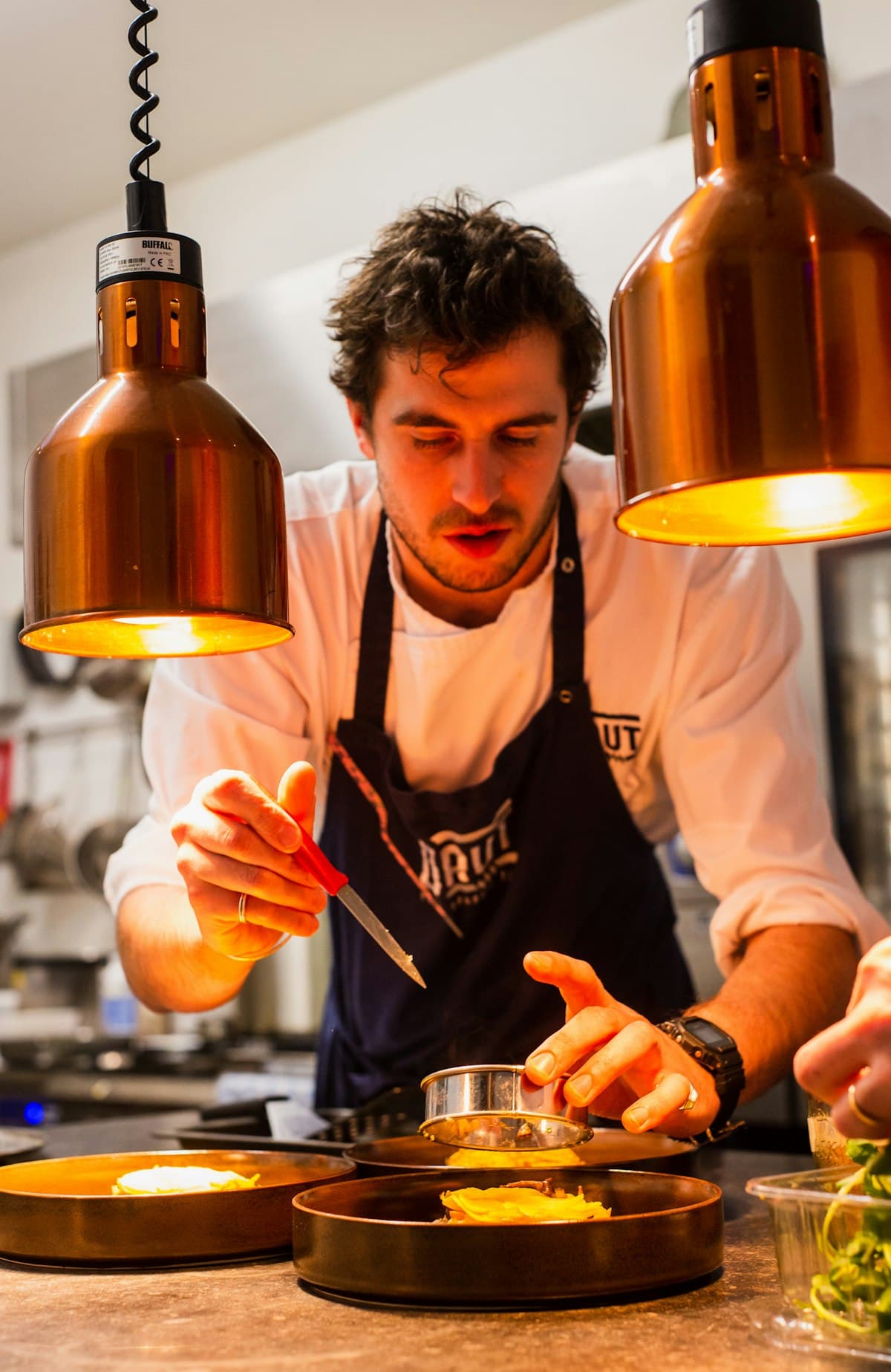 Chef plating a dish under warm copper heat lamps in a restaurant kitchen