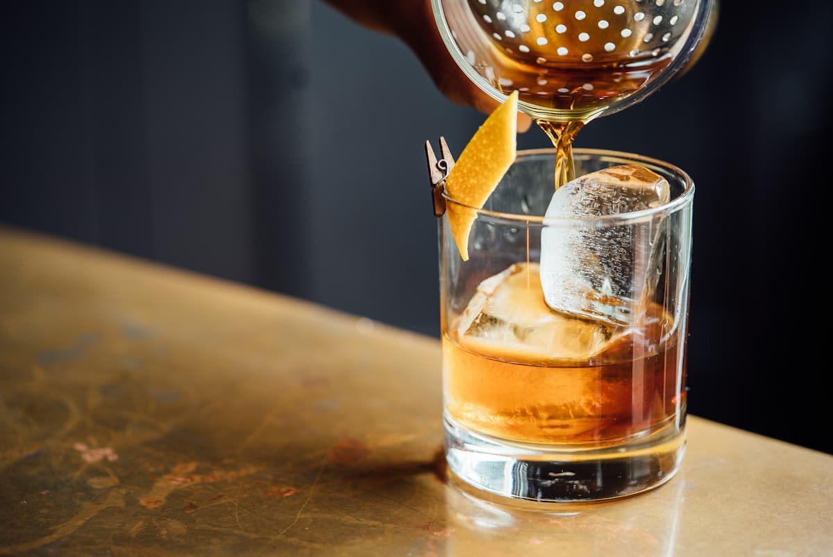 An old fashioned cocktail being strained over a large ice cube in a rocks glass on a brass bar top