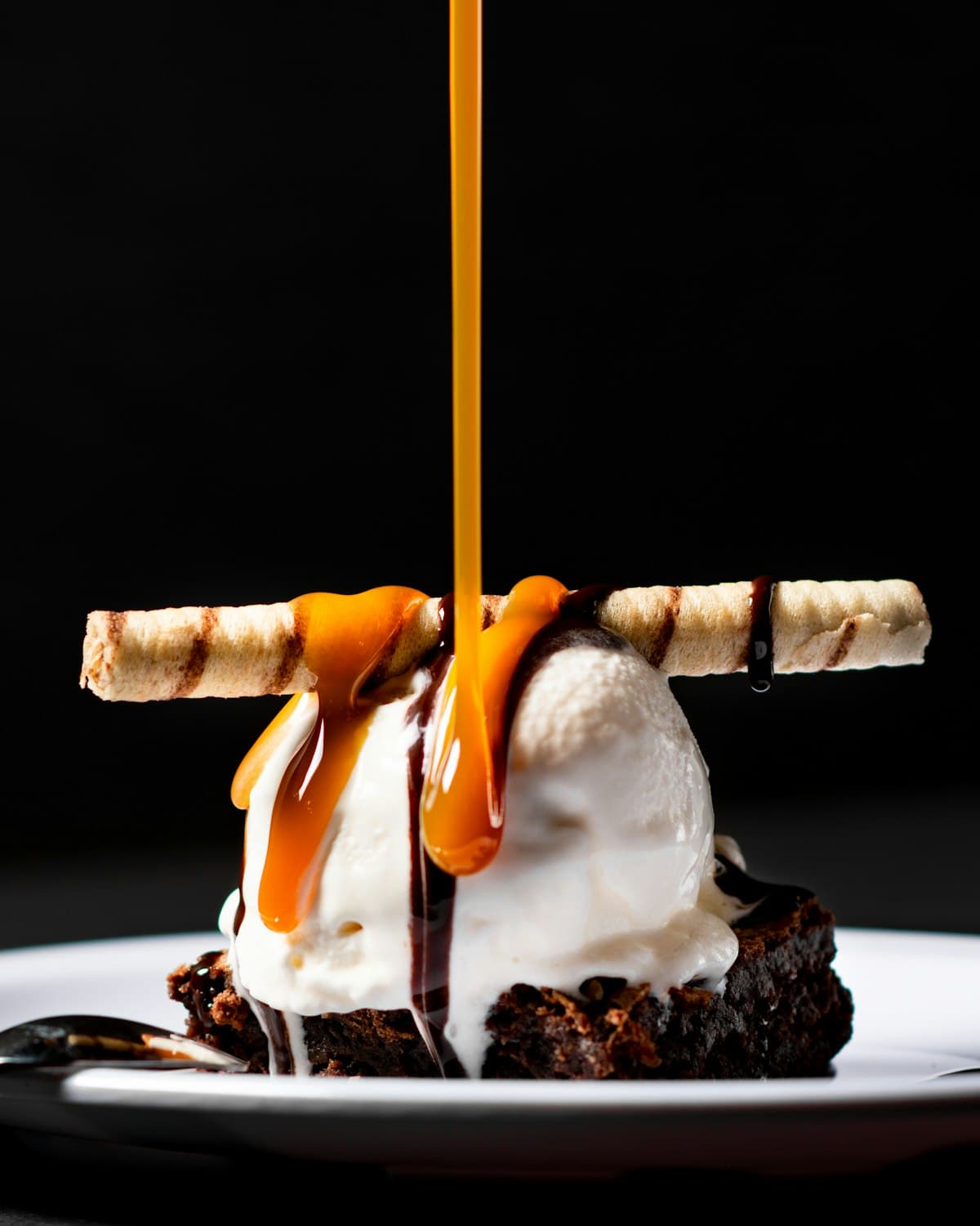 Caramel being poured over a brownie sundae with vanilla ice cream against a black background